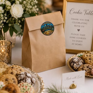 Op de afbeelding: Een bruine papieren zak verzegeld met een ronde sticker met een illustratie van de skyline van Pittsburgh en de tekst Pittsburgh Cookie Table. De zak staat bij een ingelijst bord met de tekst Cookie Table, Thank you for celebrating with us.