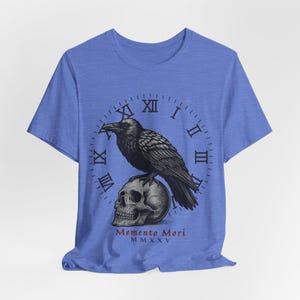 May include: Blue t-shirt featuring a black raven perched atop a skull. A clock face with Roman numerals surrounds the raven and skull. The words "Memento Mori" and the Roman numeral "MMXXV" are printed below the skull.