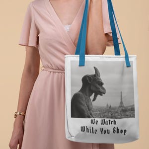 Op de afbeelding: Witte draagtas met blauwe handvatten. De tas toont een zwart-wit afbeelding van een waterspuwer met uitzicht op een stadslandschap met de Eiffeltoren. De tekst "We Watch While You Shop" staat eronder.