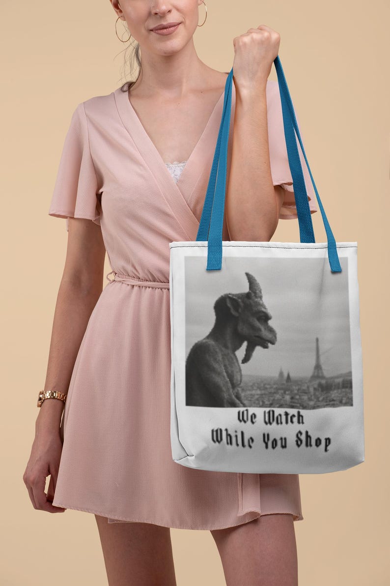 Op de afbeelding: Witte draagtas met blauwe handvatten. De tas toont een zwart-wit afbeelding van een waterspuwer met uitzicht op een stadslandschap met de Eiffeltoren. De tekst "We Watch While You Shop" staat eronder.