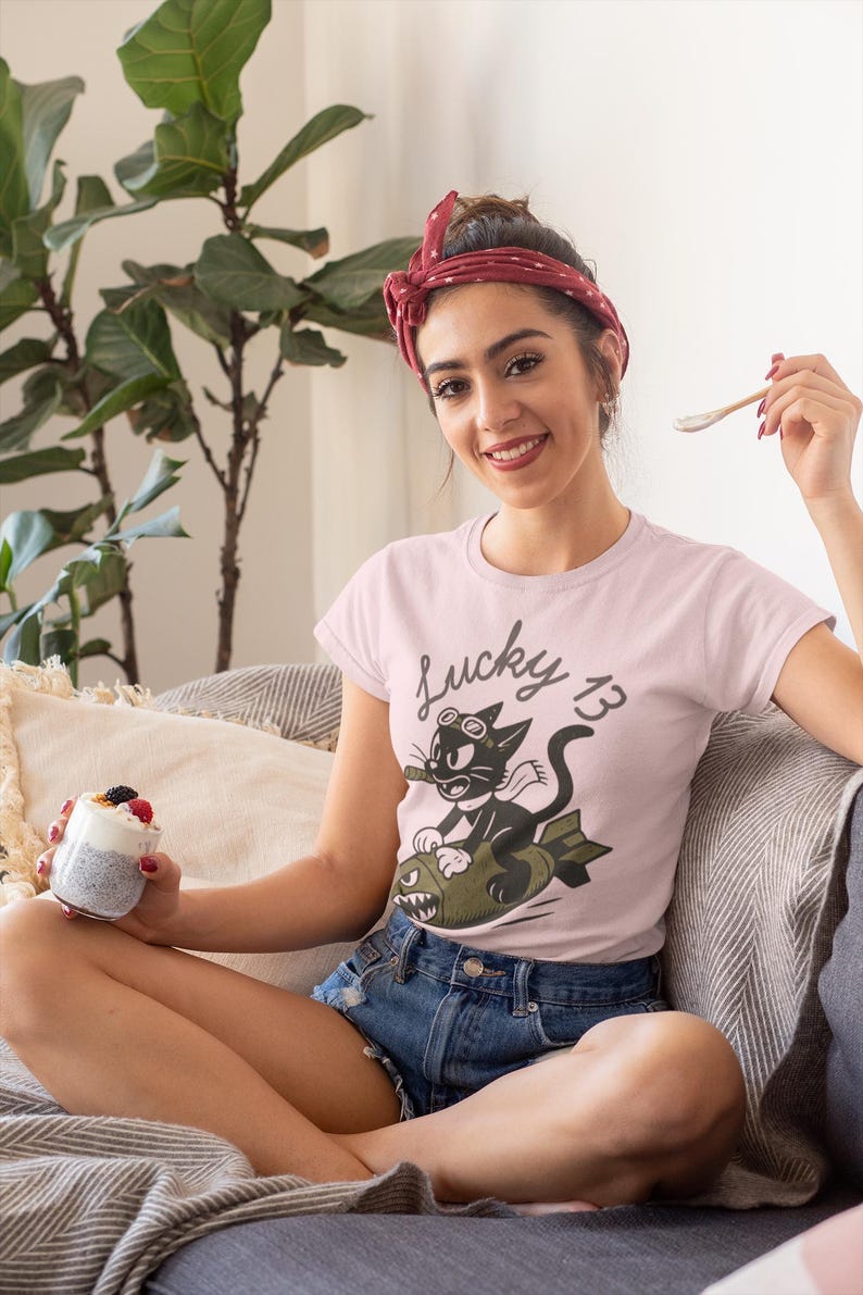 May include: A light pink short-sleeved t-shirt featuring a graphic of a black cartoon cat wearing goggles and a scarf while riding a green bomb. The text "Lucky 13" is printed in a cursive font above the cat illustration on the front of the shirt.