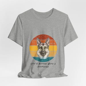 May include: Light blue t-shirt featuring a German Shepherd dog graphic within a retro-style circular design. The graphic includes a dog's face against a backdrop of orange, yellow, and blue horizontal stripes. Below the graphic, the text reads 'Life's Better with a Shepherd'.