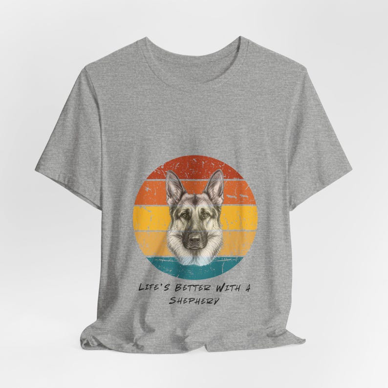 May include: Light blue t-shirt featuring a German Shepherd dog graphic within a retro-style circular design. The graphic includes a dog's face against a backdrop of orange, yellow, and blue horizontal stripes. Below the graphic, the text reads 'Life's Better with a Shepherd'.