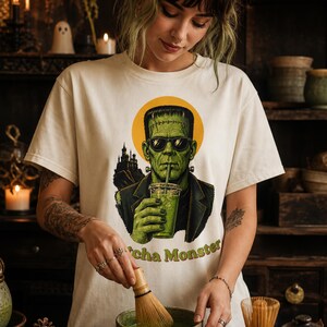 Może przedstawiać: Kremowy t-shirt z grafiką przedstawiającą zielonego potwora w okularach przeciwsłonecznych, pijącego z przezroczystego kubka. Wz&oacute;r zawiera sylwetkę ciemnego zamku, ż&oacute;łte koło w tle oraz zielony napis Matcha Monster.
