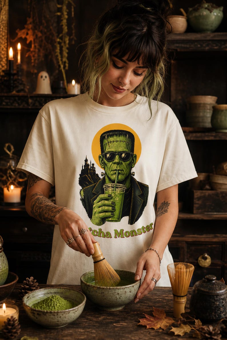Może przedstawiać: Kremowy t-shirt z grafiką przedstawiającą zielonego potwora w okularach przeciwsłonecznych, pijącego z przezroczystego kubka. Wz&oacute;r zawiera sylwetkę ciemnego zamku, ż&oacute;łte koło w tle oraz zielony napis Matcha Monster.