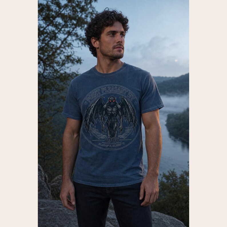 May include: A blue short-sleeved t-shirt featuring a circular graphic of a winged creature with glowing red eyes. The text reads Point Pleasant WV and 1966. The design includes a dark, detailed illustration of a moth-like figure with large wings.