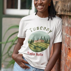 May include: A light-colored t-shirt with a circular graphic depicting a Tuscan landscape, including rolling hills, trees, and buildings. The text "Tuscany" arches above the image, with "Italia Bella" below. The person is wearing blue jeans.