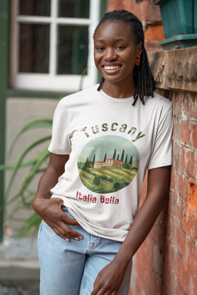 May include: A light-colored t-shirt with a circular graphic depicting a Tuscan landscape, including rolling hills, trees, and buildings. The text "Tuscany" arches above the image, with "Italia Bella" below. The person is wearing blue jeans.