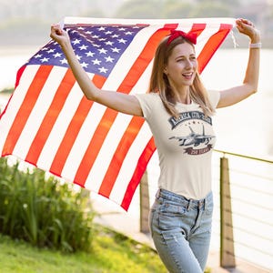 May include: A cream-colored short-sleeve t-shirt featuring a graphic of a fighter jet with the text America 1776-2026 and 250 Years. The shirt is paired with blue denim jeans and a red headband, while a large American flag is held overhead.