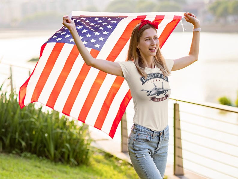 May include: A cream-colored short-sleeve t-shirt featuring a graphic of a fighter jet with the text America 1776-2026 and 250 Years. The shirt is paired with blue denim jeans and a red headband, while a large American flag is held overhead.