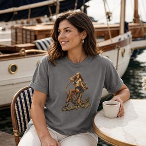 May include: A charcoal gray short-sleeve t-shirt featuring a graphic print of a pin-up style figure sitting on a large ship anchor. The text "Anchors Aweigh" is printed in a stylized font beneath the illustration on the front of the garment.