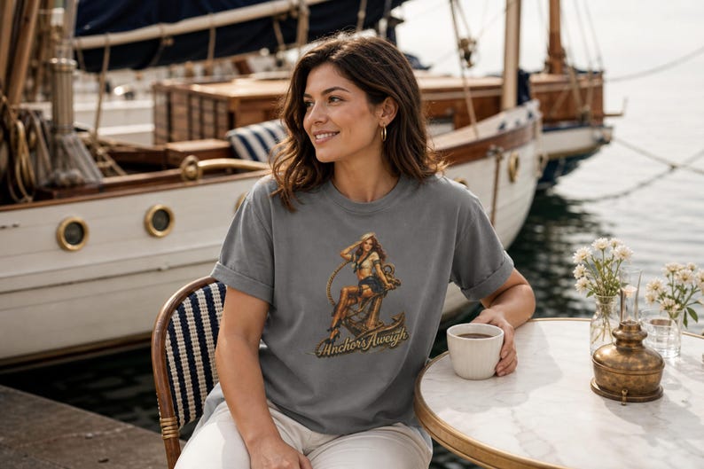 May include: A charcoal gray short-sleeve t-shirt featuring a graphic print of a pin-up style figure sitting on a large ship anchor. The text "Anchors Aweigh" is printed in a stylized font beneath the illustration on the front of the garment.