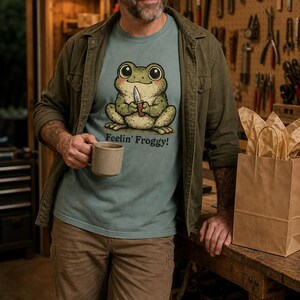 Może przedstawiać: Zakurzony niebieski t-shirt z grafiką przedstawiającą zieloną żabę trzymającą mały n&oacute;ż, z nadrukowanym poniżej tekstem Feelin' Froggy!. Żaba ma duże ciemne oczy i jasny brzuch, wykonana w stylu rysunkowym.