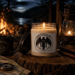 May include: A clear glass candle jar with a white label featuring a black circular graphic of a winged creature, the text Point Pleasant WV, 1966, and coordinates. The candle has a lit wick and sits next to its black metal lid on a wooden surface.
