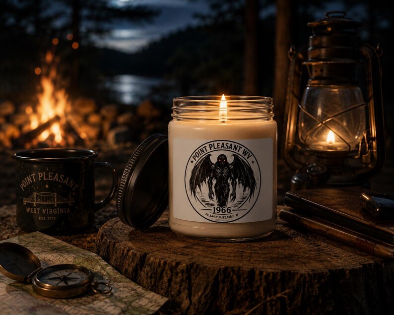 May include: A clear glass candle jar with a white label featuring a black circular graphic of a winged creature, the text Point Pleasant WV, 1966, and coordinates. The candle has a lit wick and sits next to its black metal lid on a wooden surface.