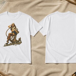 May include: A white short-sleeved t-shirt featuring a graphic print of a pin-up style sailor sitting on a large metal anchor. The text "Anchors Aweigh" is printed in a stylized font below the anchor. The back of the t-shirt is plain white.