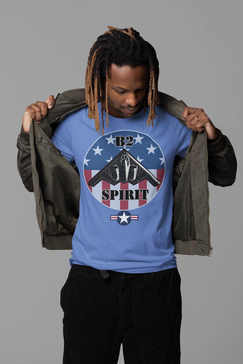 May include: A light blue t-shirt featuring a graphic of a B2 Spirit stealth bomber aircraft. The design includes a circular emblem with a blue background, white stars, and red and white stripes. The word 'SPIRIT' is printed below the aircraft, and 'B2' is at the top.