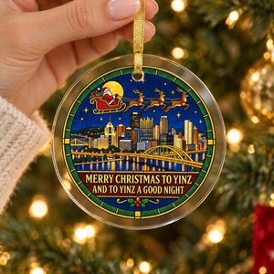 May include: A round, clear acrylic Christmas ornament featuring a stained-glass style illustration of the Pittsburgh skyline at night, with Santa and his reindeer flying above. The text reads, Merry Christmas to Yinz and to Yinz a good night.