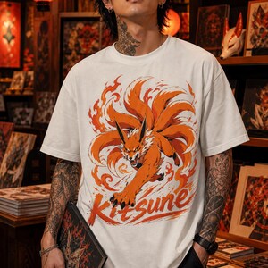 K&ouml;nnte beinhalten: Ein wei&szlig;es T-Shirt mit kurzen &Auml;rmeln und einem Grafikdruck eines orangefarbenen neunschw&auml;nzigen Fuchses, der von stilisierten Flammen umgeben ist. Das Wort Kitsune ist in orangefarbenen Buchstaben unten auf dem Grafikdesign aufgedruckt.