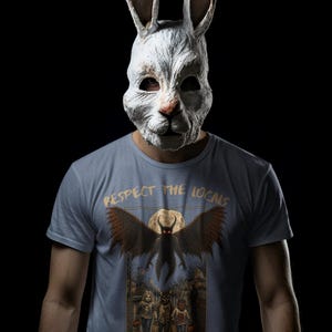 May include: A light blue t-shirt featuring a graphic print with the text "RESPECT THE LOCALS" above an illustration of a winged creature flying over three children in costumes on a street at night, worn with a weathered white rabbit mask.