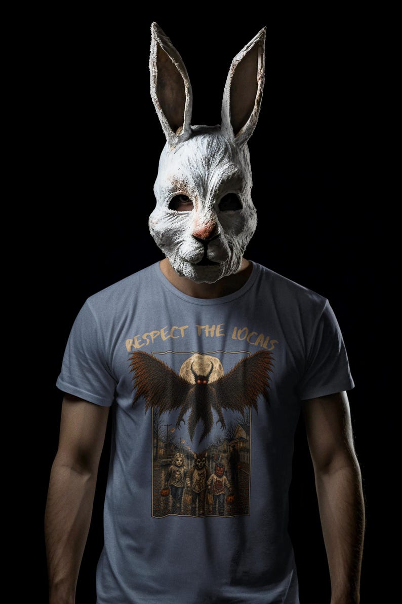 May include: A light blue t-shirt featuring a graphic print with the text "RESPECT THE LOCALS" above an illustration of a winged creature flying over three children in costumes on a street at night, worn with a weathered white rabbit mask.