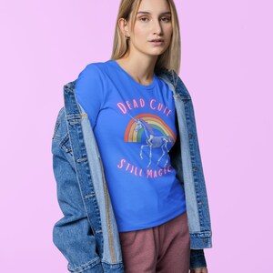 May include: A blue t-shirt with the text "DEAD CUTE STILL MAGIC" and a graphic of a unicorn skeleton over a rainbow. The shirt is worn with a denim jacket and pink pants.