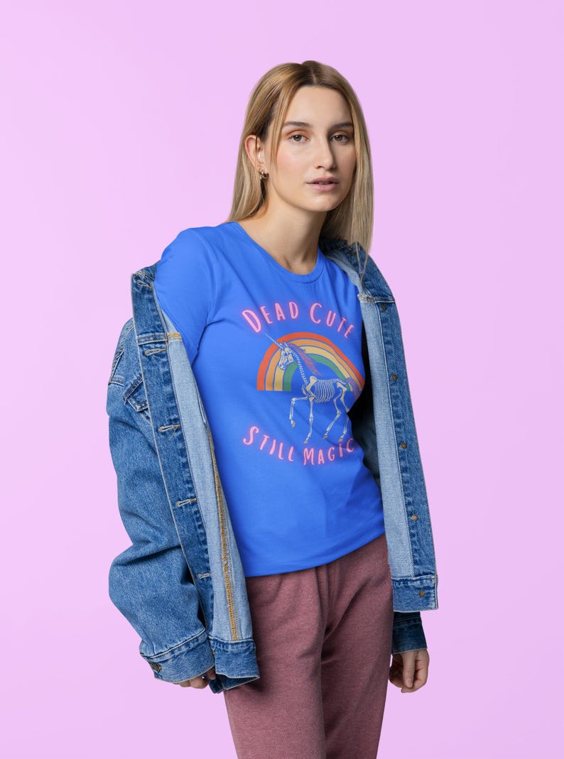 May include: A blue t-shirt with the text "DEAD CUTE STILL MAGIC" and a graphic of a unicorn skeleton over a rainbow. The shirt is worn with a denim jacket and pink pants.