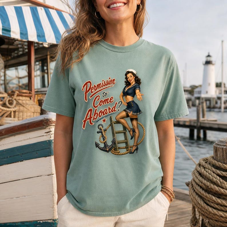 Op de afbeelding: Een saliegroen t-shirt met korte mouwen en een grafische print van een pin-up zeeman die naast een ladder en een anker staat. De tekst "Permission to Come Aboard?" is in rode cursieve letters boven de illustratie van de zeeman gedrukt.