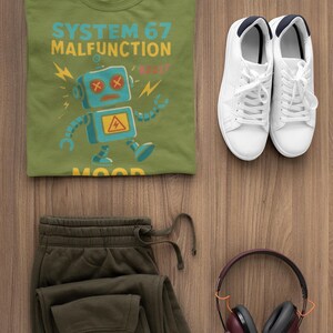 May include: Olive green t-shirt with a blue robot graphic and the text "SYSTEM 67 MALFUNCTION". Paired with olive green sweatpants, white sneakers, and maroon headphones on a wooden surface. A casual outfit.