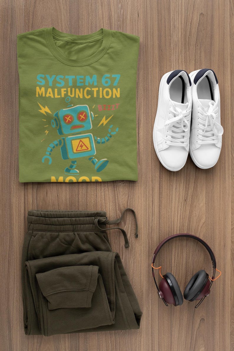 May include: Olive green t-shirt with a blue robot graphic and the text "SYSTEM 67 MALFUNCTION". Paired with olive green sweatpants, white sneakers, and maroon headphones on a wooden surface. A casual outfit.