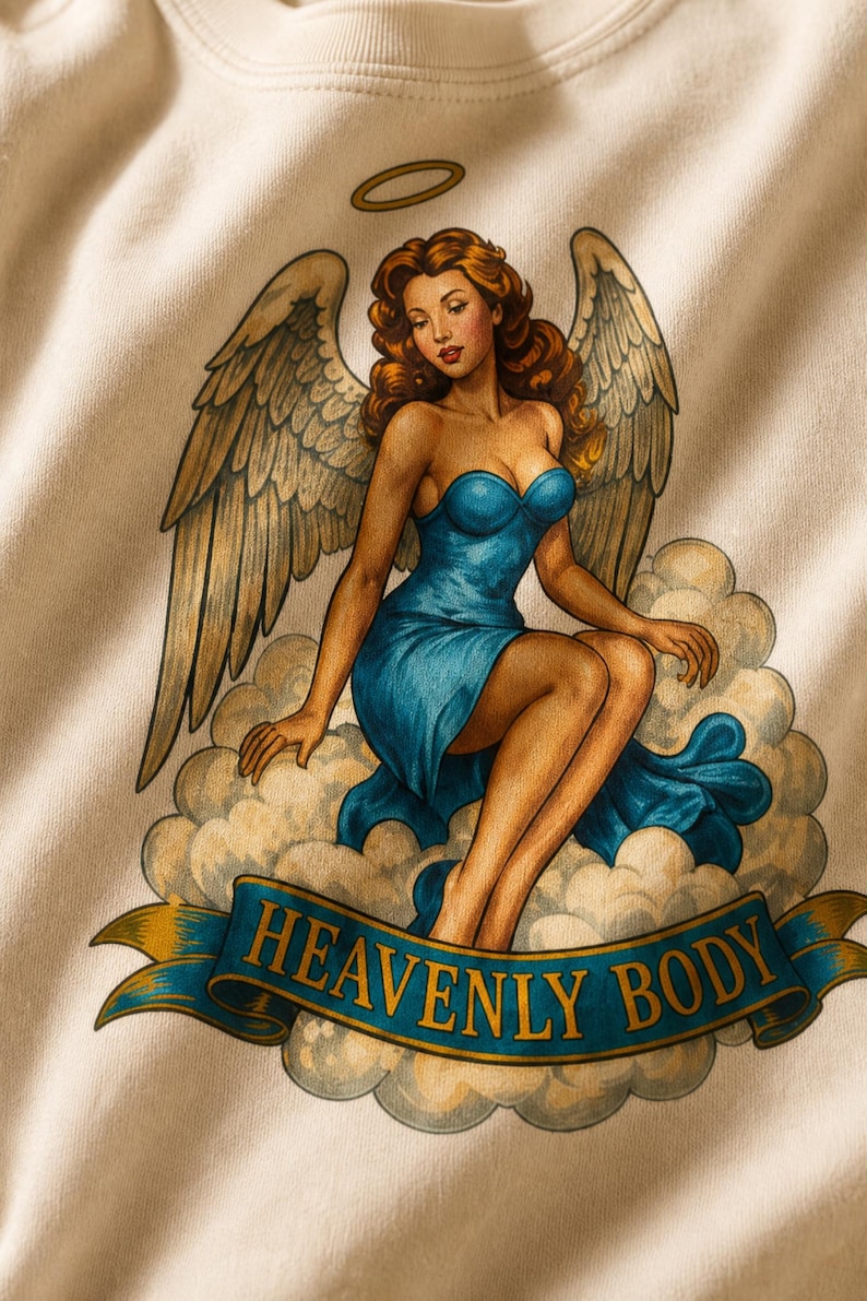 May include: A cream-colored sweatshirt featuring a graphic print of an angel with auburn hair, large wings, and a halo, wearing a blue strapless dress while sitting on clouds. A blue banner at the bottom displays the text HEAVENLY BODY in gold.