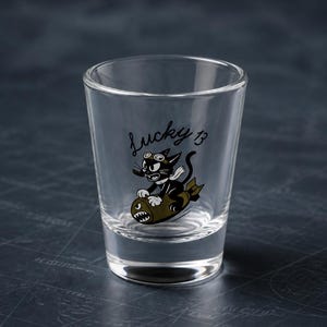 May include: A clear glass shot glass featuring a black cartoon cat wearing goggles and a white scarf, riding an olive green bomb. The text "Lucky 13" is printed in black script above the cat. The glass has a thick base and tapered sides.