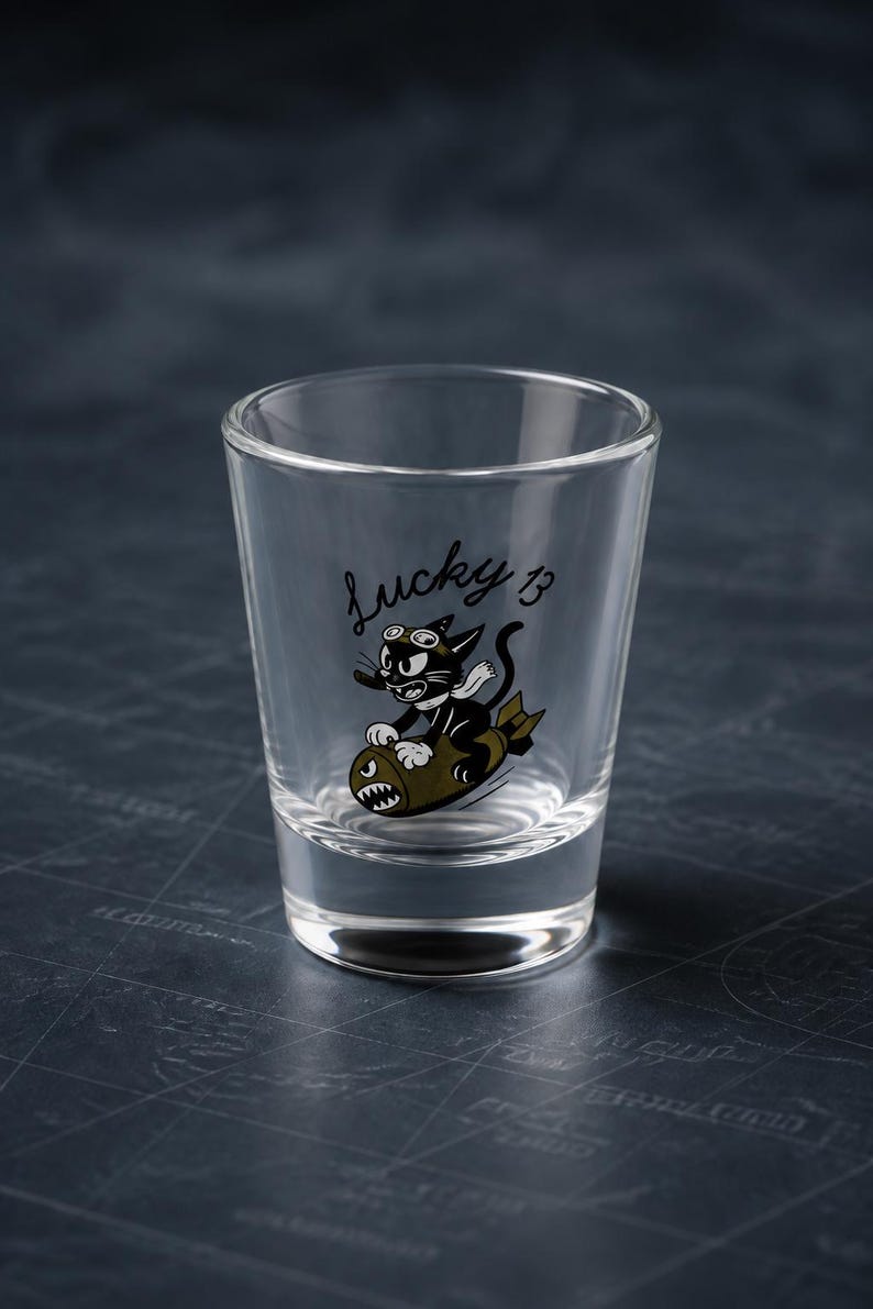 May include: A clear glass shot glass featuring a black cartoon cat wearing goggles and a white scarf, riding an olive green bomb. The text "Lucky 13" is printed in black script above the cat. The glass has a thick base and tapered sides.