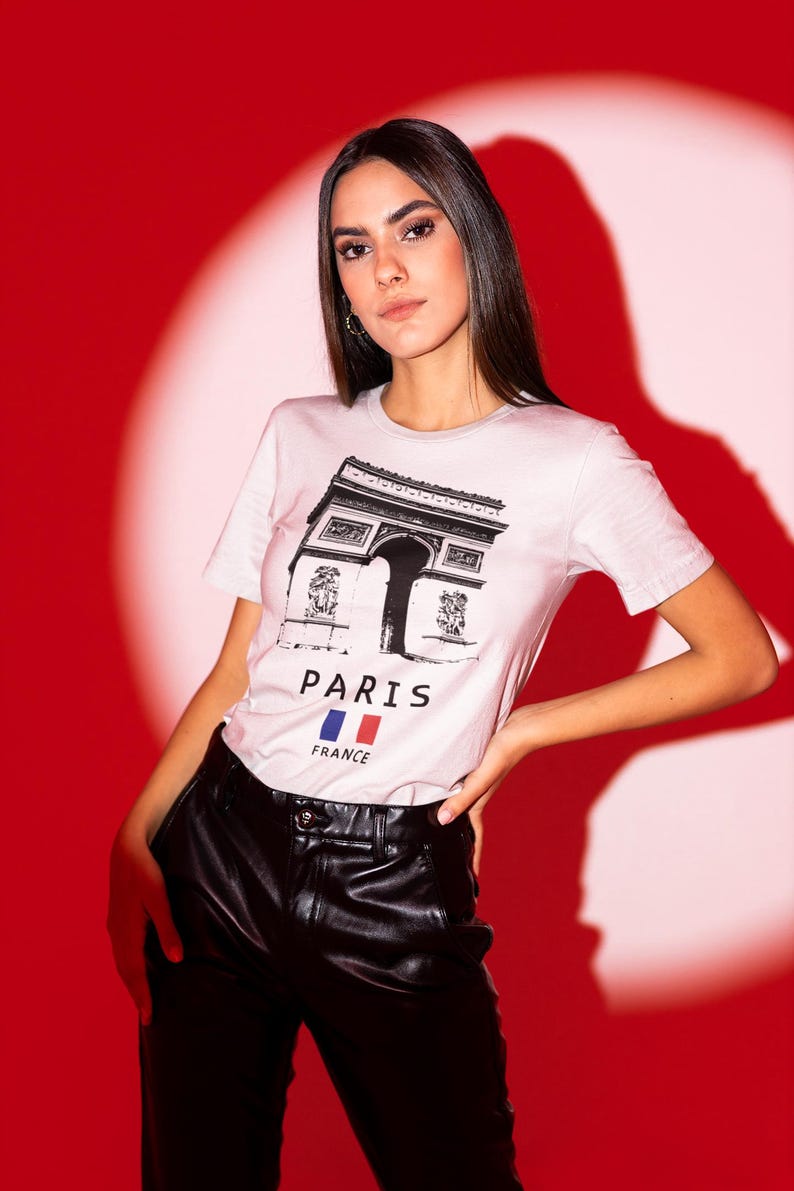 Puede incluir: Camiseta rosa claro con un dibujo en blanco y negro del Arco del Triunfo. Las palabras "PARIS" y "FRANCE" est&aacute;n impresas debajo, junto con la bandera francesa. La persona lleva pantalones de cuero negro.