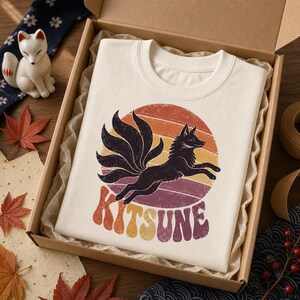 May include: A cream-colored t-shirt featuring a graphic of a black nine-tailed fox leaping in front of a distressed, multi-colored sunset circle. Below the fox, the word KITSUNE is printed in a bold, retro-style font with orange and purple tones.