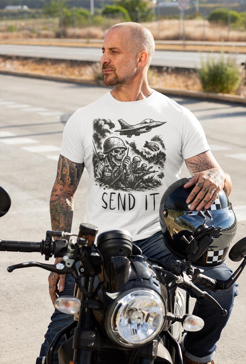 May include: White t-shirt with a black graphic of a skull soldier, fighter jets, and the text "SEND IT." The shirt is worn by a person sitting on a black motorcycle, holding a black helmet. The motorcycle has a round headlight.