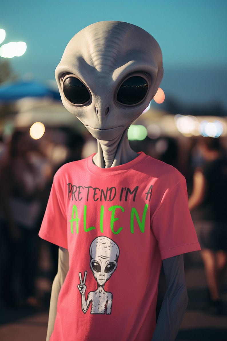 May include: A gray alien with large black eyes wearing a pink t-shirt. The shirt has the text "PRETEND I'M A ALIEN" in green and white, along with a cartoon alien giving a peace sign.