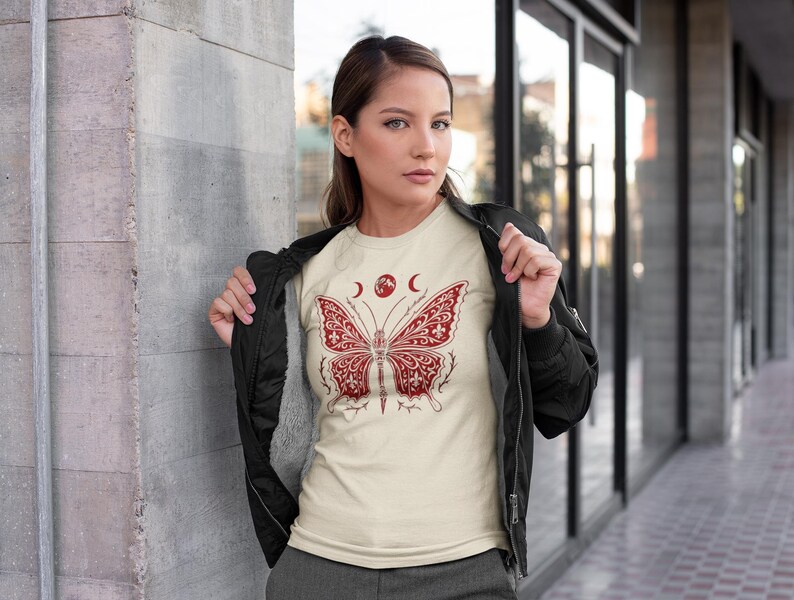 May include: A cream-colored t-shirt featuring a red butterfly design. The butterfly has intricate patterns and is positioned below two crescent moons and a circular shape. The person is wearing a black jacket over the t-shirt, standing in front of a building with glass windows.
