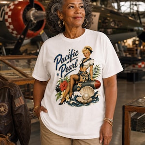 May include: A white t-shirt featuring a graphic print of a pin-up style character sitting on a bomb, with the text Pacific Pearl, a large open oyster shell with a pearl, tropical flowers, palm leaves, and ocean waves in blue, red, and green.