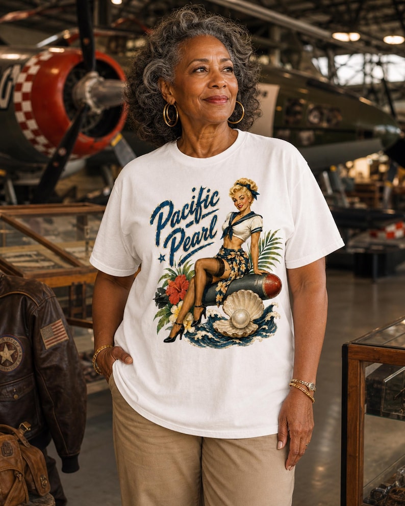 May include: A white t-shirt featuring a graphic print of a pin-up style character sitting on a bomb, with the text Pacific Pearl, a large open oyster shell with a pearl, tropical flowers, palm leaves, and ocean waves in blue, red, and green.