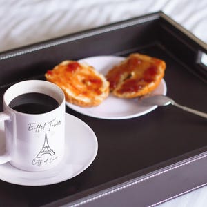 May include: A white mug with black coffee and a saucer, featuring an Eiffel Tower design and the text "City of Love." Two slices of toast with jam sit on a white plate, with a spoon. The items are on a dark brown tray.