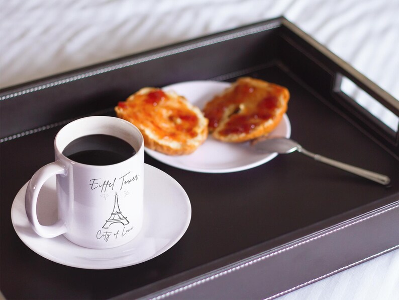 May include: A white mug with black coffee and a saucer, featuring an Eiffel Tower design and the text "City of Love." Two slices of toast with jam sit on a white plate, with a spoon. The items are on a dark brown tray.