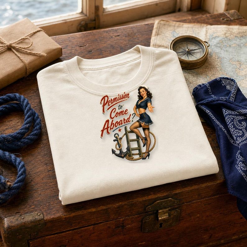 Op de afbeelding: Een cr&egrave;mekleurig t-shirt met een afbeelding van een pin-up zeeman die op een ladder naast een anker staat. De tekst luidt Permission to Come Aboard? in rode letters. Het ontwerp bevat nautische elementen zoals een touw.