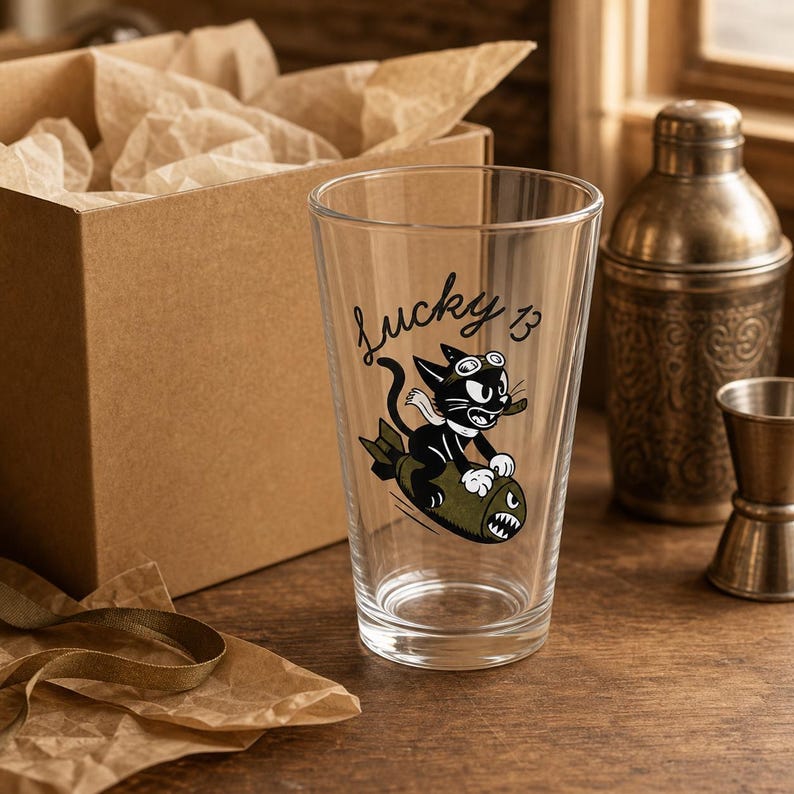 May include: A clear glass pint tumbler featuring a black and white graphic of a cartoon cat wearing goggles and a scarf while riding an olive green bomb. The text "lucky 13" is printed in black script above the cat illustration on the glass surface.