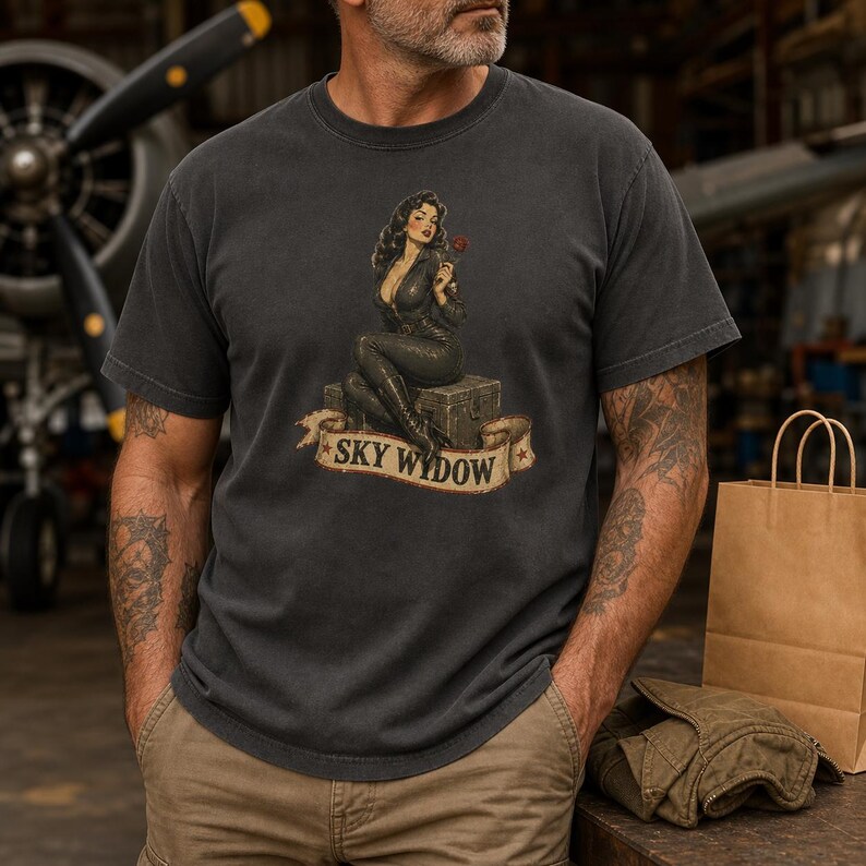 May include: A charcoal gray short-sleeve t-shirt featuring a graphic print of a woman in a black jumpsuit sitting on a crate, holding a red rose. Below the figure is a beige banner with the text SKY WYDOW in black capital letters.
