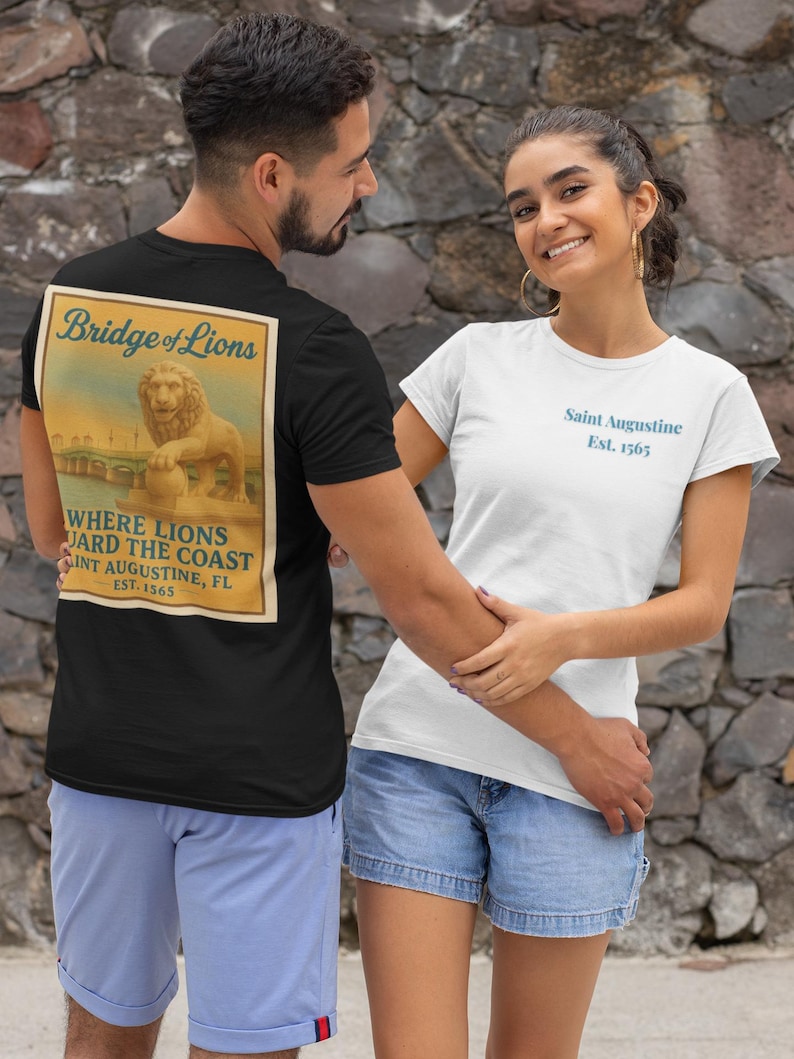 Może przedstawiać: Czarny t-shirt z grafiką w stylu vintage na plecach z napisem "Bridge of Lions" i "Where Lions Guard the Coast, Saint Augustine, FL, Est. 1565." Widoczny jest r&oacute;wnież biały t-shirt z napisem "Saint Augustine Est. 1565" w kolorze niebieskim.