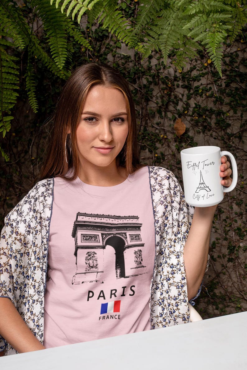 May include: A woman wearing a pink t-shirt with a black graphic of the Arc de Triomphe and the word "PARIS" below it. She is also holding a white mug with the Eiffel Tower and the words "City of Love" printed on it. She is wearing a floral patterned kimono.