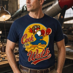 May include: A navy blue short-sleeve t-shirt featuring a graphic of a cartoon rooster wearing a brown aviator helmet and red boxing gloves. The rooster is centered within a yellow circle, with the text Cock of the Walk printed in red.