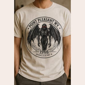 May include: A cream-colored short-sleeve t-shirt featuring a circular black graphic of a winged creature with glowing red eyes. The text reads Point Pleasant WV, 1966, and the coordinates 38.8443 N, 82.1385 W within the circular design.