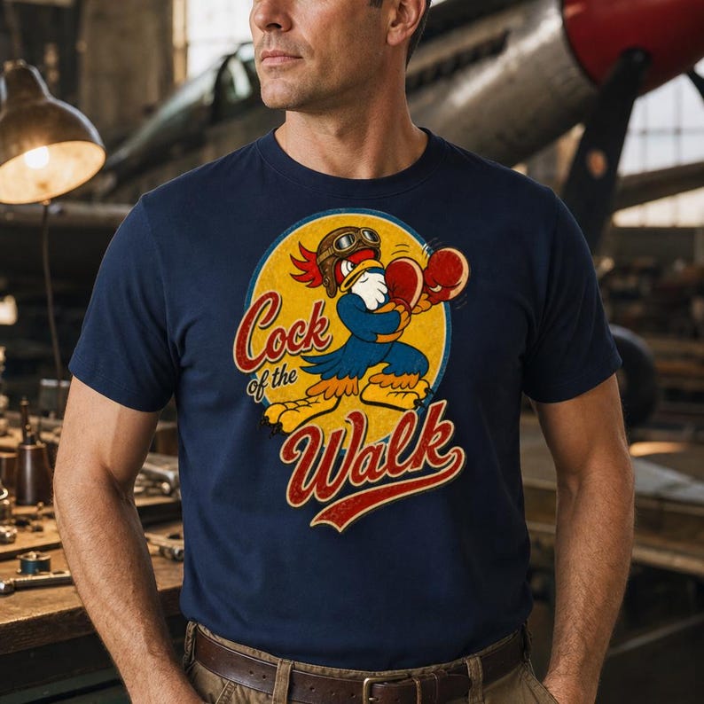 May include: A navy blue t-shirt featuring a graphic of a cartoon rooster wearing aviator goggles and red boxing gloves. The rooster is positioned inside a yellow circle with the text Cock of the Walk written in a red, stylized cursive font.